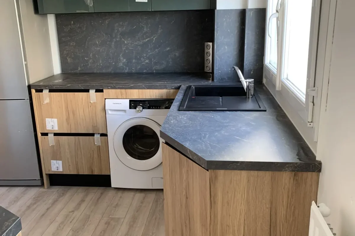 cuisine avec meubles bas bois clair, plan de travail marbré foncé. Elle comprend une machine à laver, un évier noir et un réfrigérateur