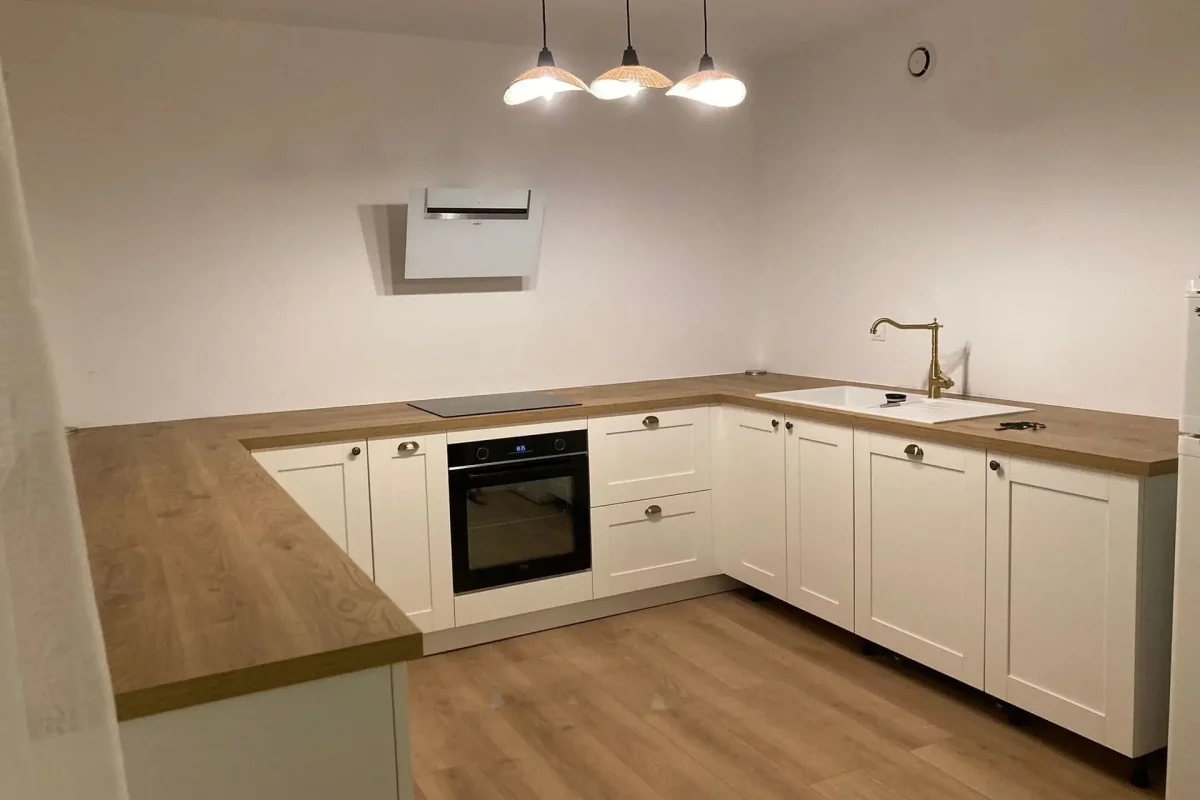 cuisine blanche épurée, avec plans de travail et sol en bois. Elle est équipée d'un robinet doré, d'une hotte murale blanche et de trois suspensions lumineuses.