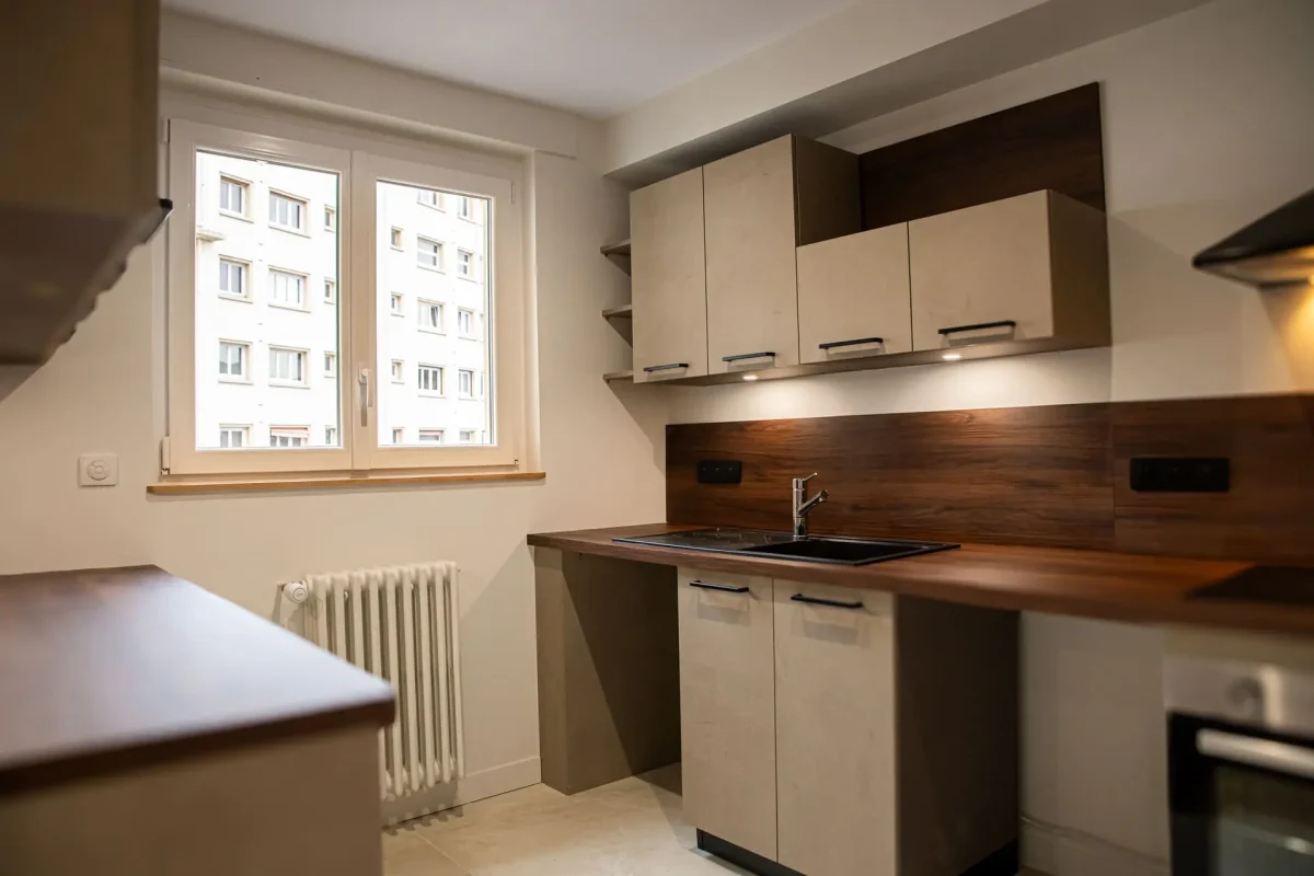 Cuisine moderne avec meubles beige sable, plan de travail en bois foncé et évier noir sous fenêtre.